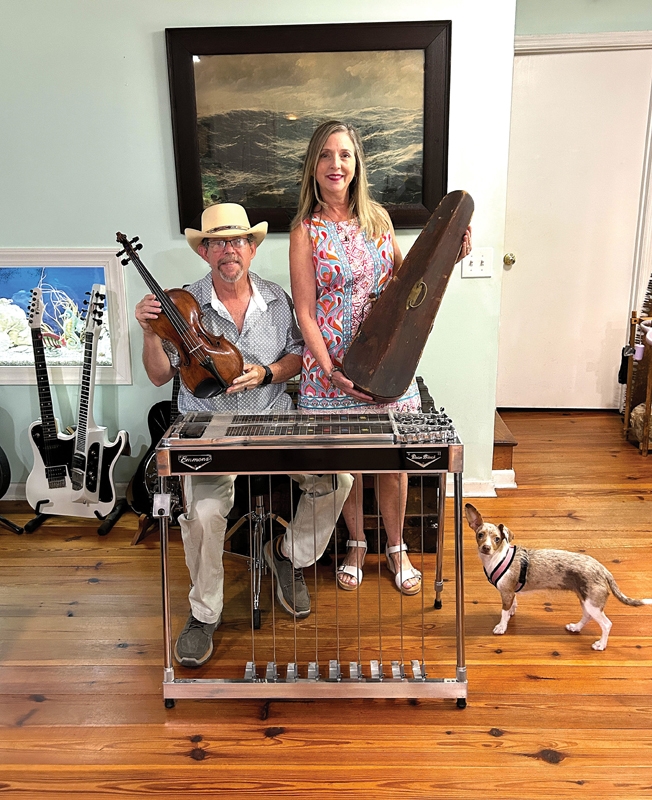 Dean and Terri Black of Pawleys Island in their home, along with their dog, Ruby, pose behind Dean’s pedal steel guitar. They’re holding his great-grandfather’s violin and its original case. A patented hybrid guitar/lap steel guitar creation of Black’s sits on a stand.